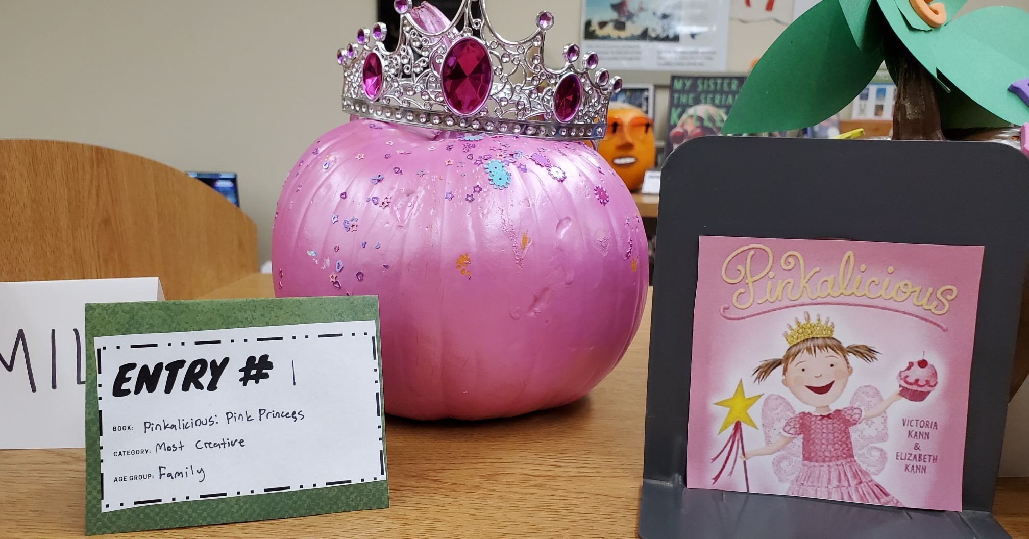 Pumpkin painted pink with a crown on it, sitting on a table next to a Pinkalicious book.