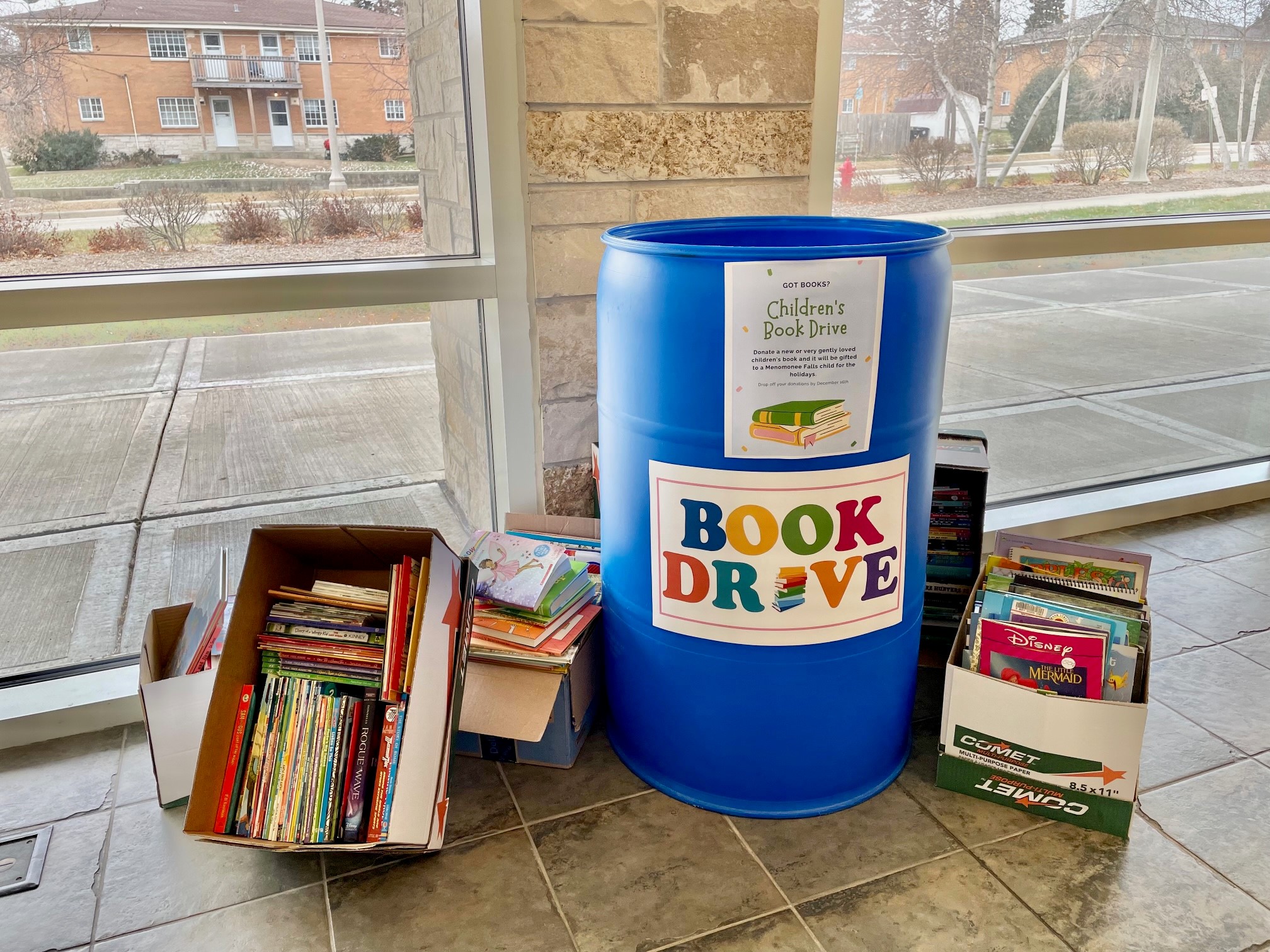Barrel with sign that says "Book Drive." Boxes of books sit next to barrel on the floor.