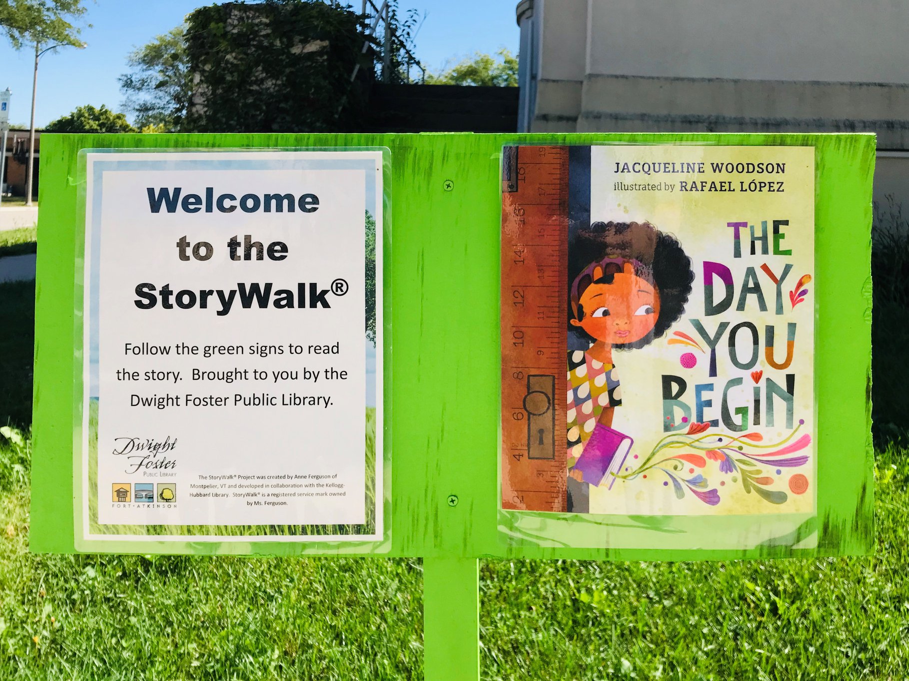 Green wooden board with title page of the book "The Day You Begin" attached to it. The board is standing outside the library. Another sign on the board says "Welcome to the StoryWalk"