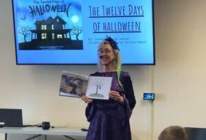 A woman reading the book "The Twelve Days of Halloween" in front of a large TV screen with the book cover on it.