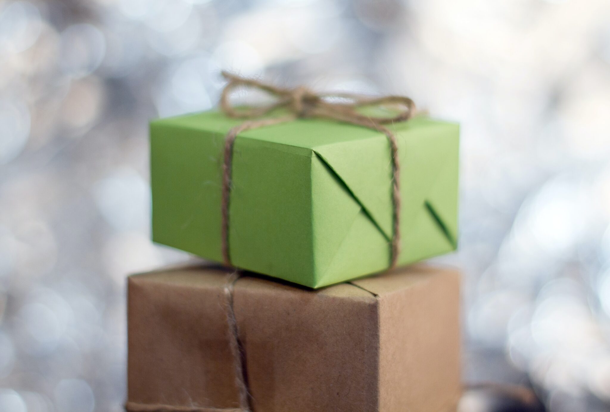 A box wrapped in green paper and tied with brown twine sits atop a box wrapped in brown paper tied with brown twine. Blurred twinkle lights are behind them.